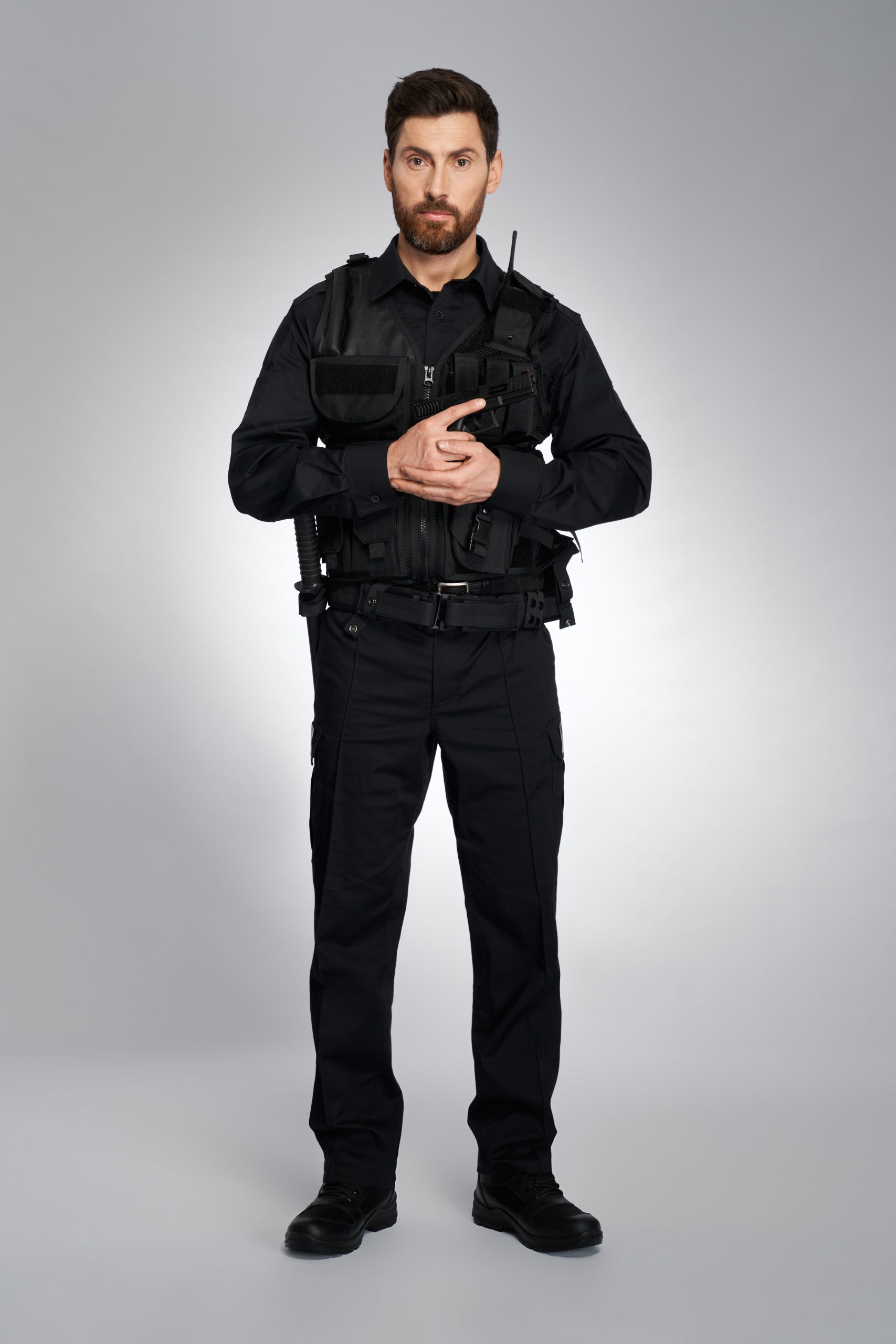 Attractive male cop with ammunition holding gun with both hands. Front view of bearded man in black body armor looking at camera, on gray studio background. Concept of work, police, rescue.